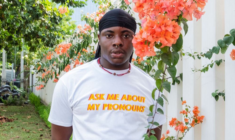 Ein junger Mann mit schwarzem Durag steht zwischen den Ästen eines lachsfarben blühenden Baumes. Die Aufschrift seines T-Shirts liest "Ask Me About My Pronouns".